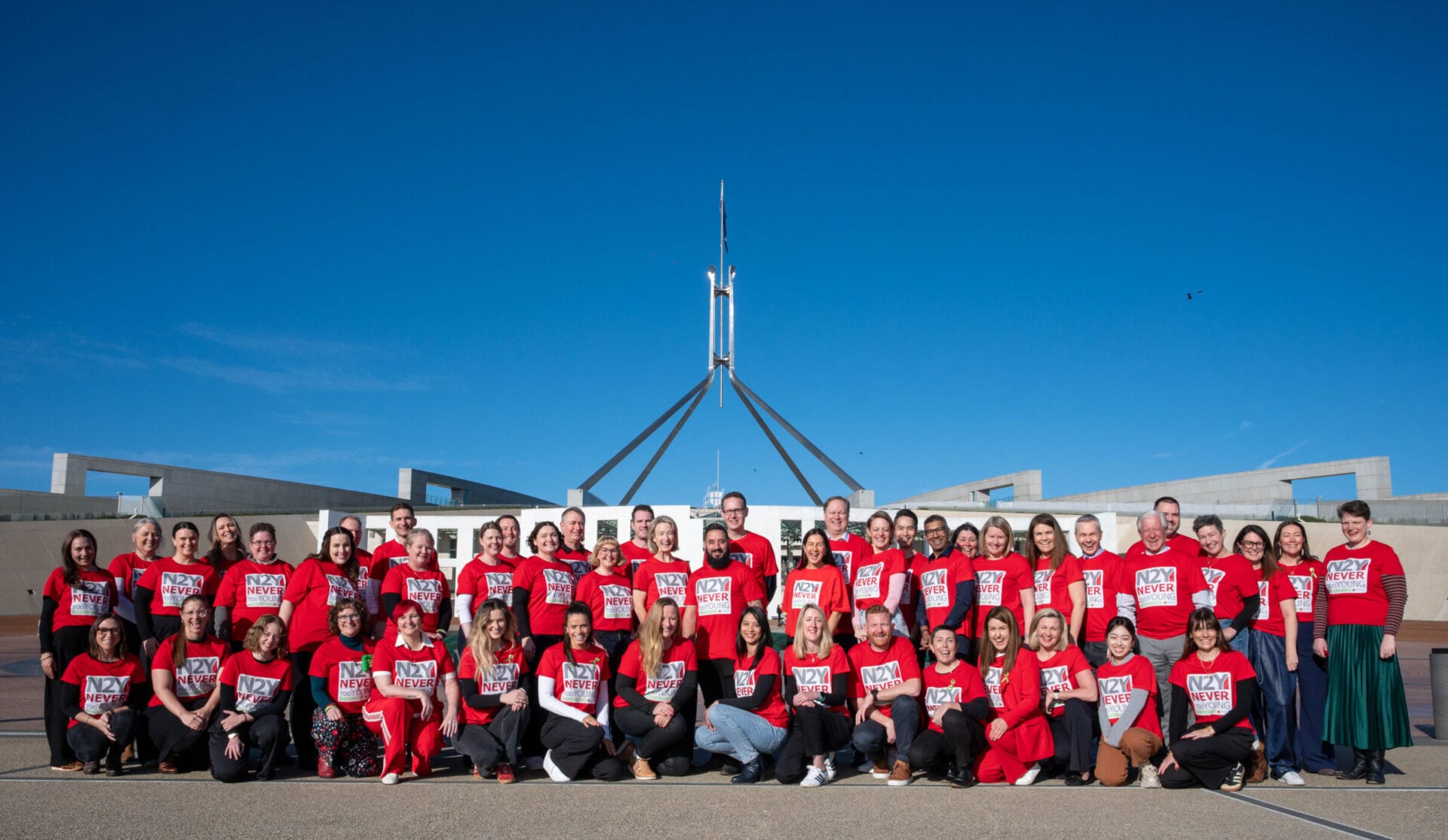 A large group of people in red shirts stands in front of a modern building under a clear blue sky.