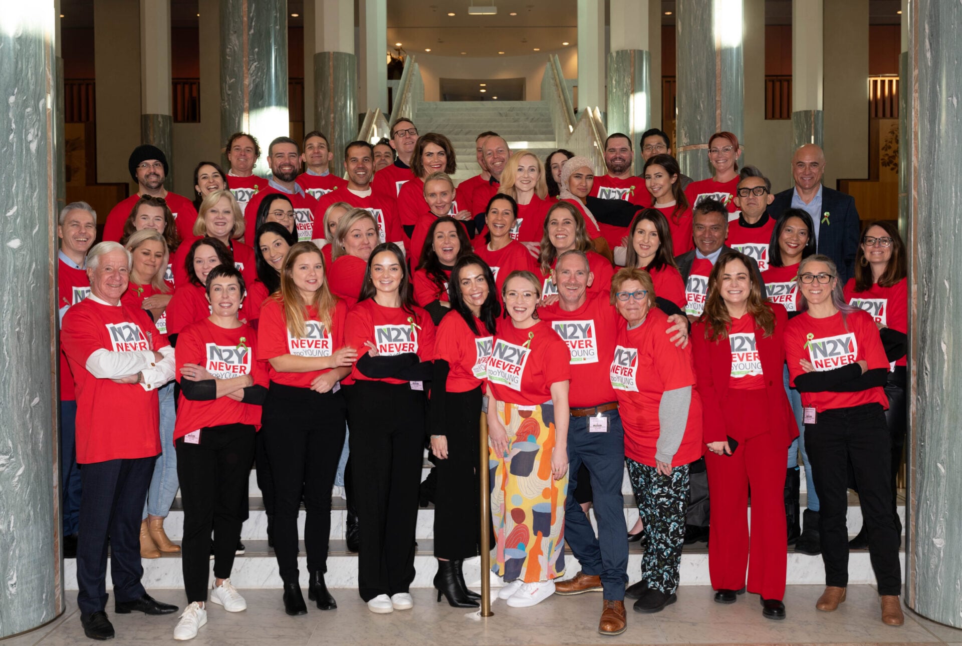A group of diverse people wearing red shirts with "N2Y NEVER TOO YOUNG" gathered on stairs in a public building.