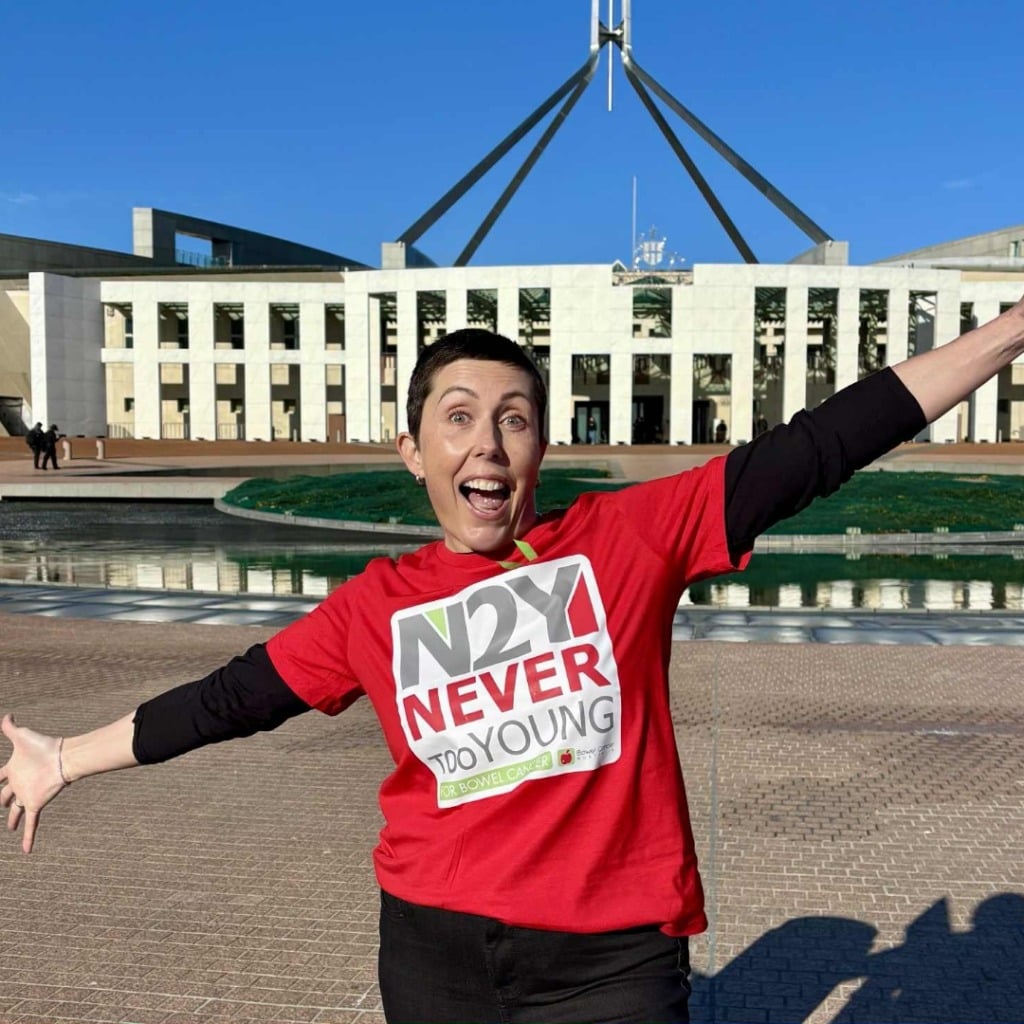 A person in a red shirt with "N2Y NEVER TOO YOUNG" stands joyfully in front of a government building.