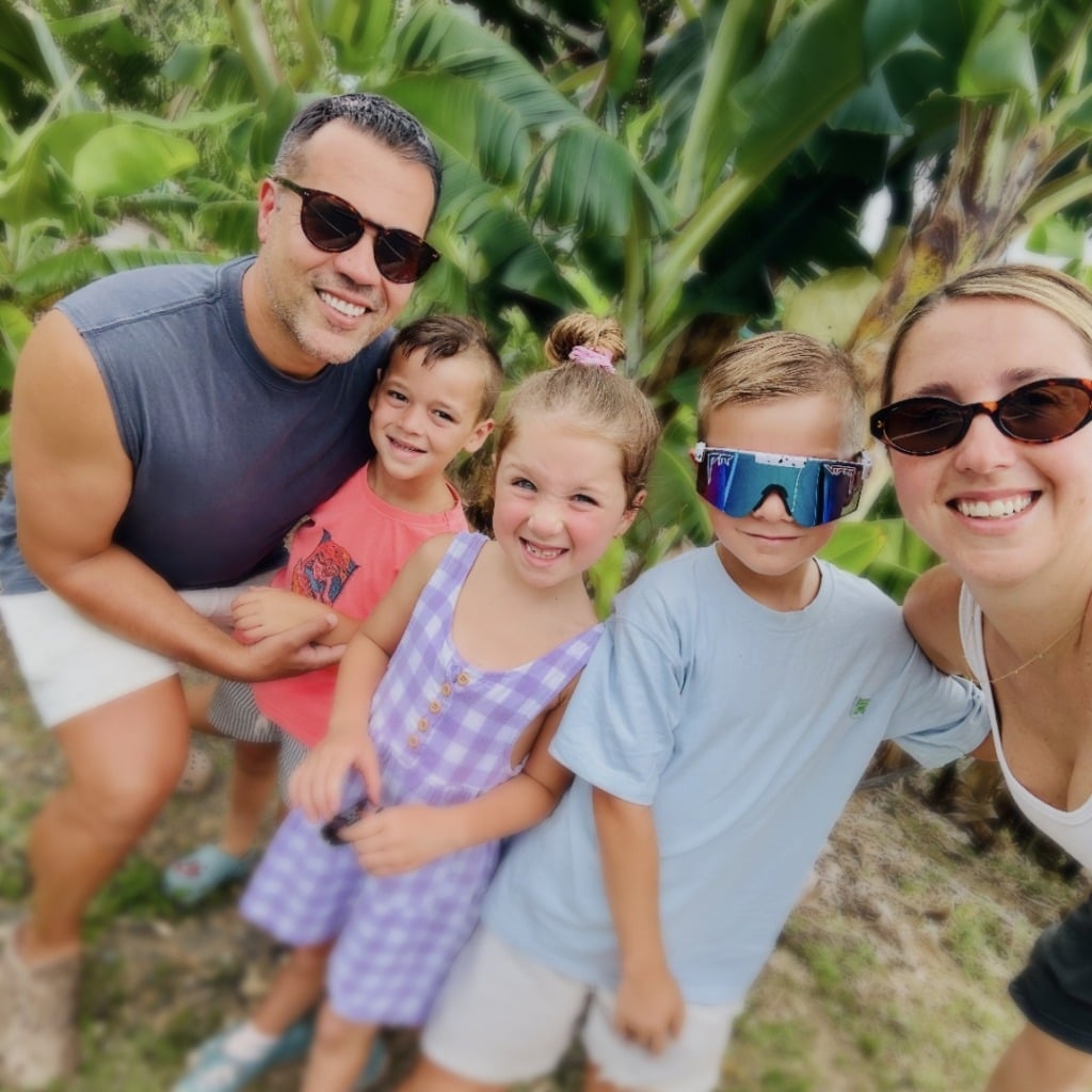 A smiling family of five poses outdoors among banana plants, wearing casual clothing and sunglasses.