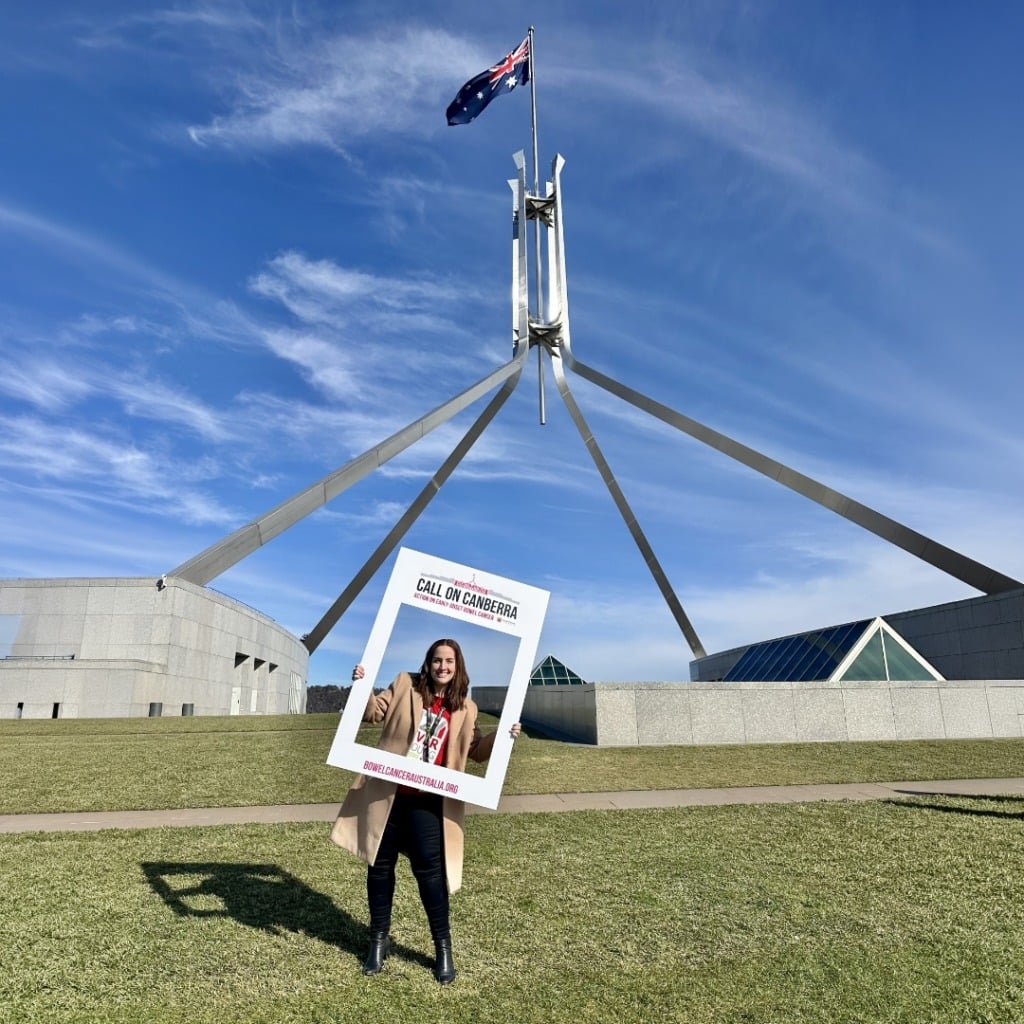 A woman holds a sign reading "CALL ON CANBERRA" in front of the Australian Parliament House under a blue sky.