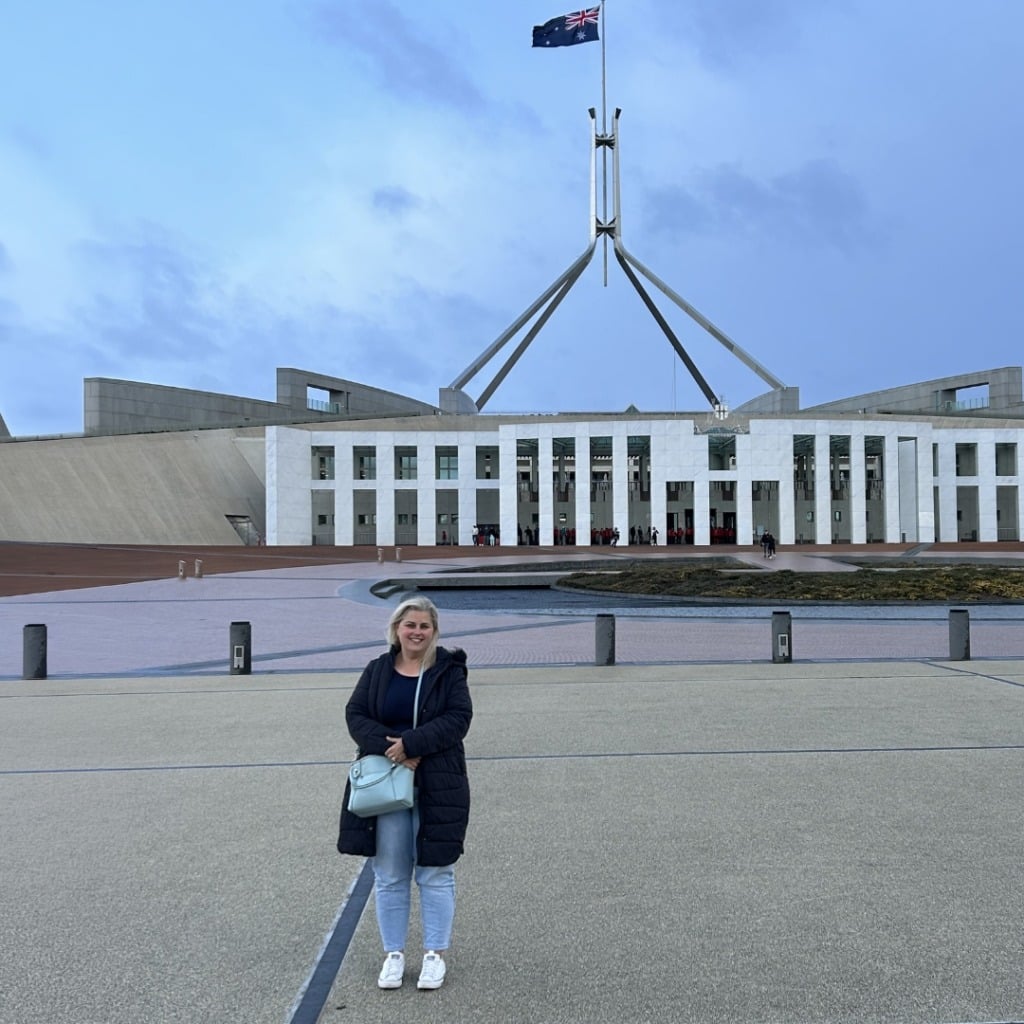 A woman stands in front of the Australian Parliament House, with a flag on a tall pole and a cloudy sky above.