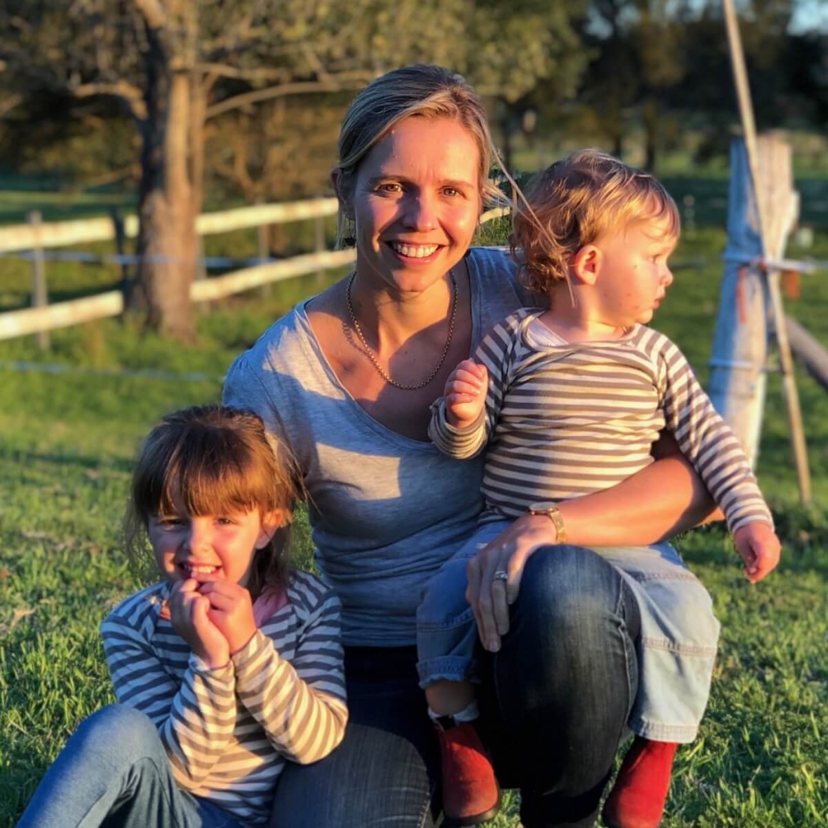 A woman sits on grass with two children, one on her lap and one beside her, all smiling in a sunny outdoor setting.