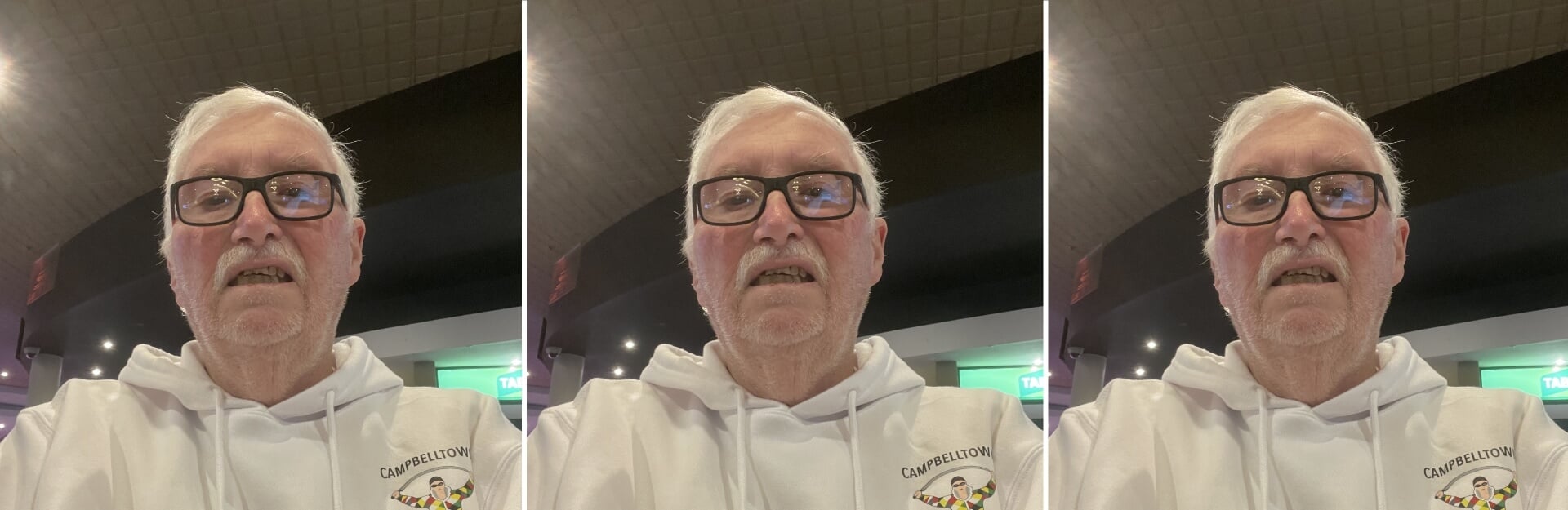 A man with gray hair and glasses wearing a white hoodie sits in a dimly lit venue, smiling at the camera.