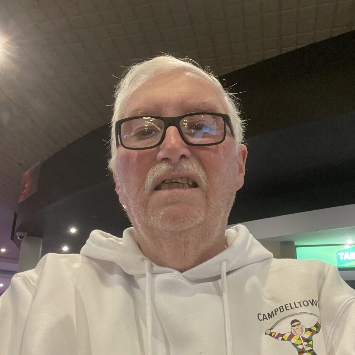 An older man with glasses and white hair, wearing a white hoodie, smiles slightly in a dimly lit venue.