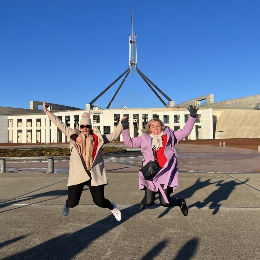 Two women jump joyfully in front of a modern building under a clear blue sky.