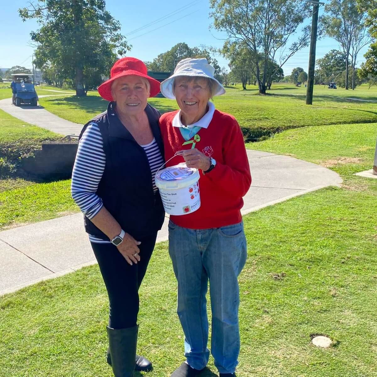 Two women stand on a grassy area, smiling. One holds a donation bucket. Both wear hats and casual clothing.