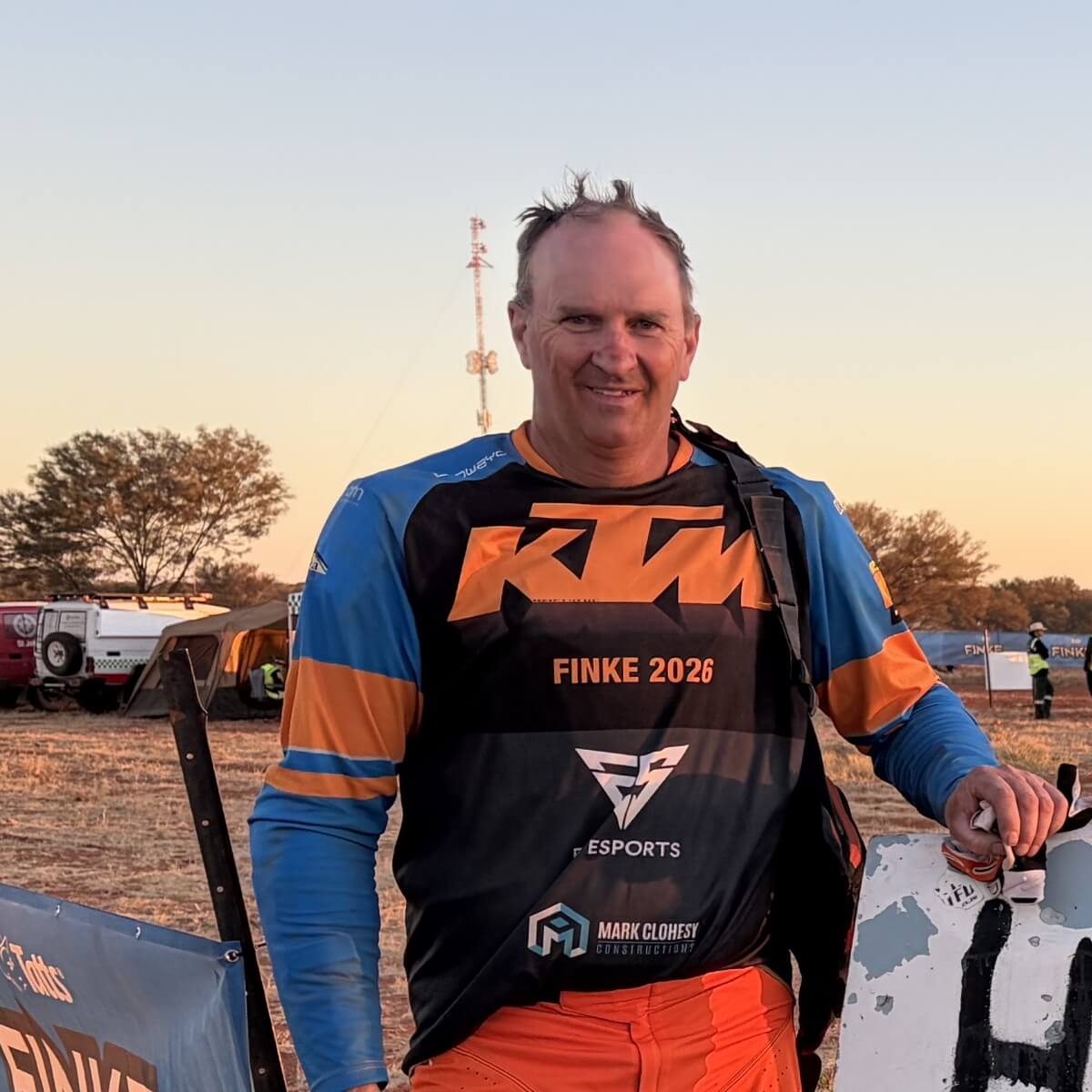 A man in an orange and black KTM shirt stands smiling, with a desert backdrop and vehicles in the distance.