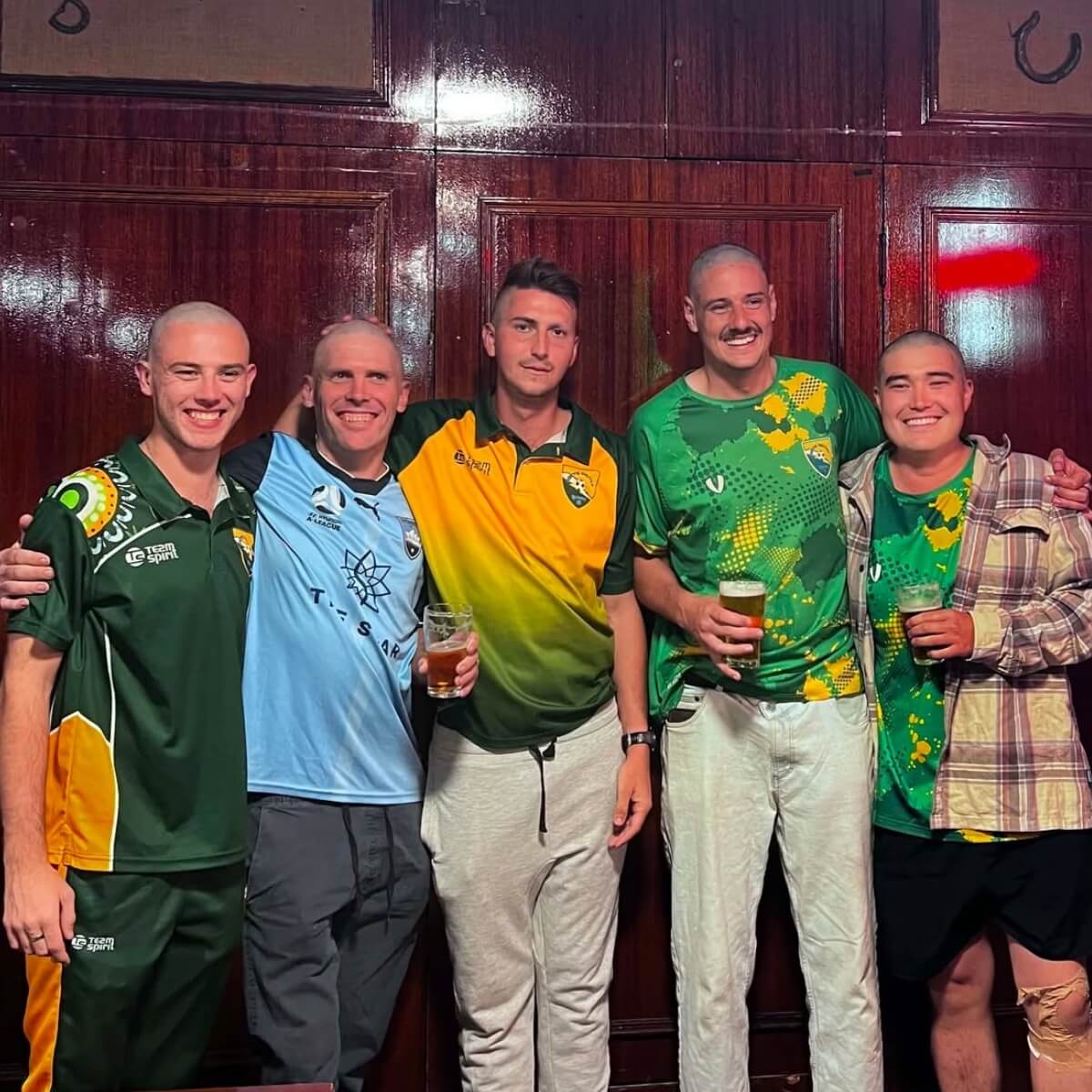 Five men with shaved heads stand together, wearing colorful sports jerseys, holding drinks in a pub setting.