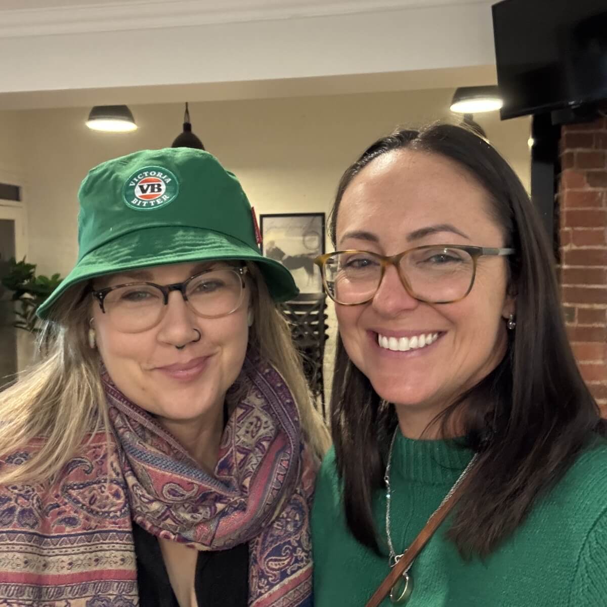 Two women smile together in a cozy indoor setting. One wears a green bucket hat and a patterned scarf.