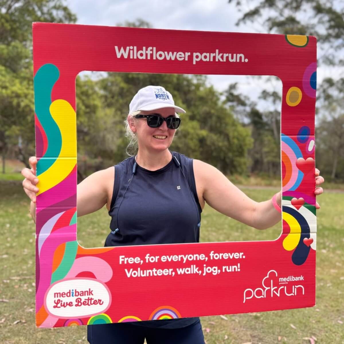 A woman in sunglasses and a cap holds a colorful frame promoting Wildflower parkrun in a grassy area.