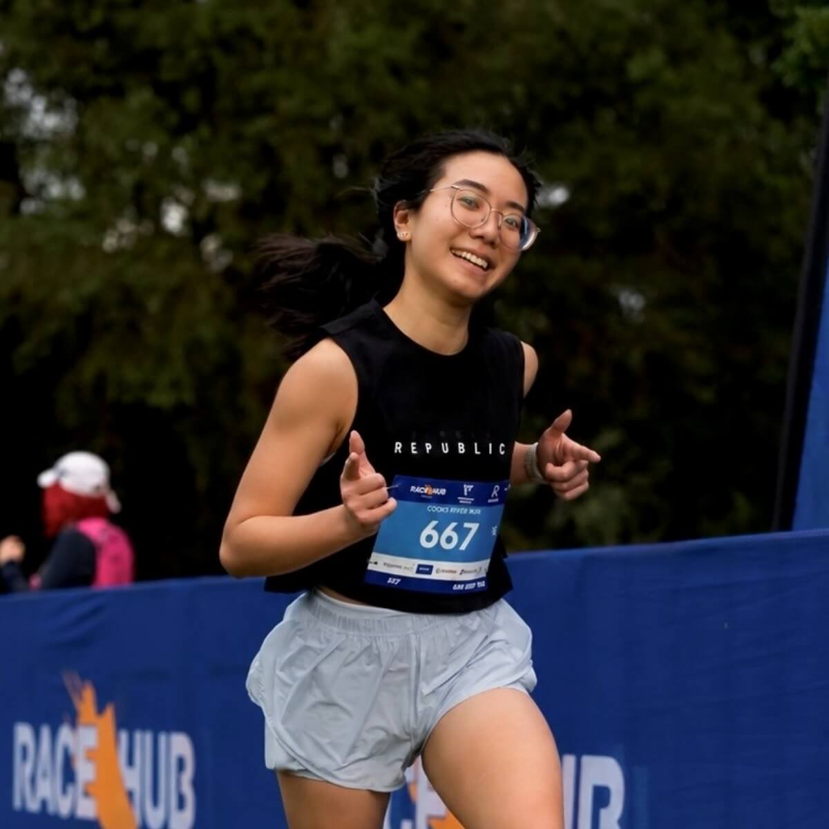 A young woman runs happily in athletic gear, wearing a bib number 667, with a blue banner in the background.