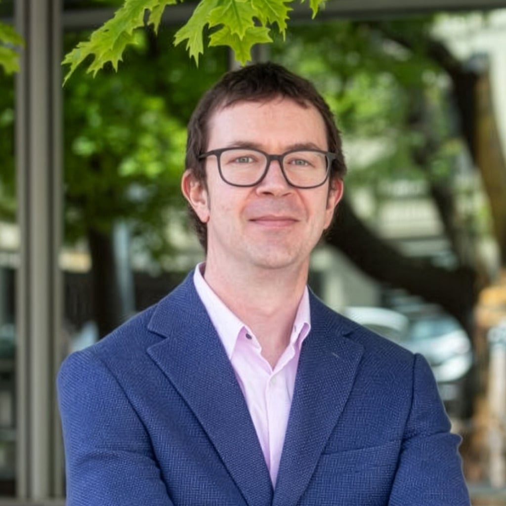 A person in a blazer stands outdoors, smiling, with trees and buildings in the background, related to early-onset bowel cancer research.