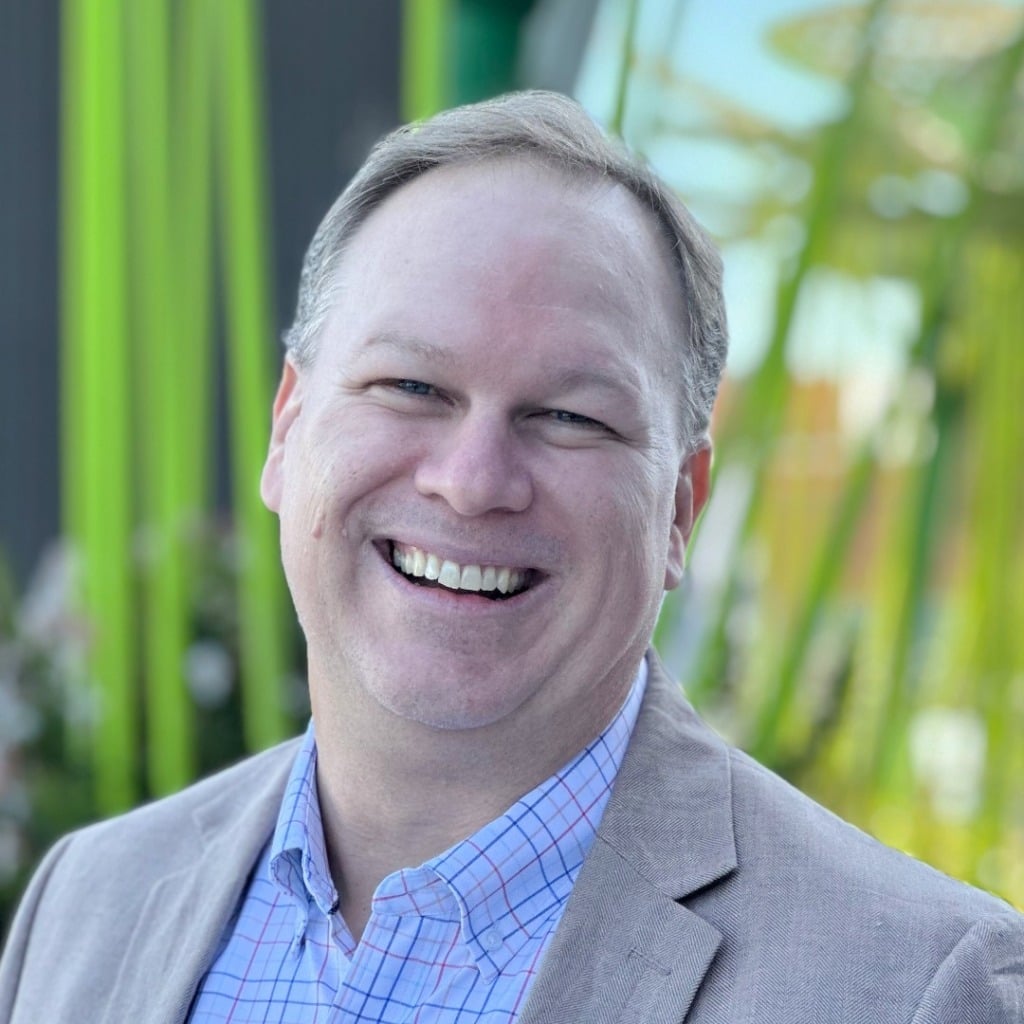 A smiling man in a light blazer stands outdoors with green plants in the background, representing an early-onset bowel cancer centre of research excellence.