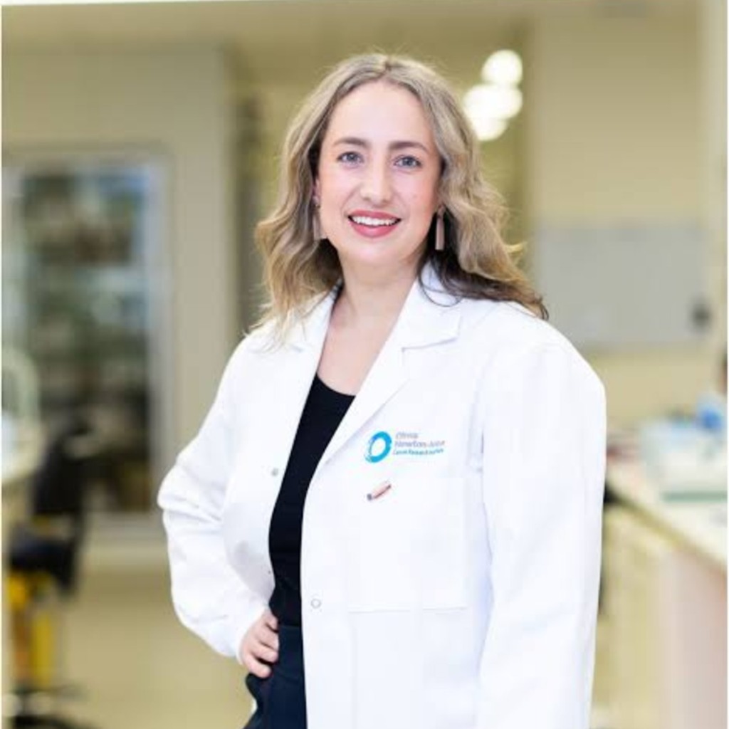 A smiling woman in a lab coat stands in a laboratory setting, representing the early-onset bowel cancer centre of research excellence.
