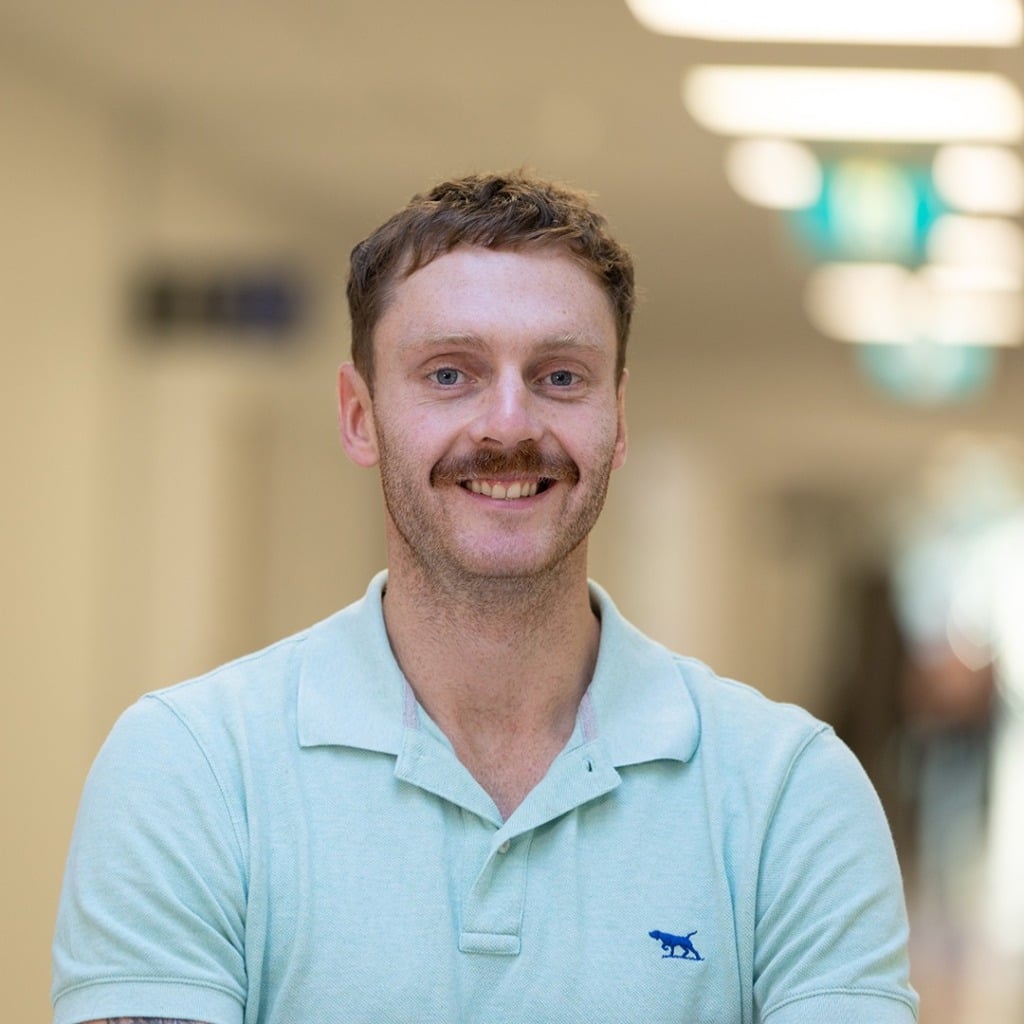 A smiling man with a mustache, wearing a light green polo shirt, stands in a well-lit corridor.