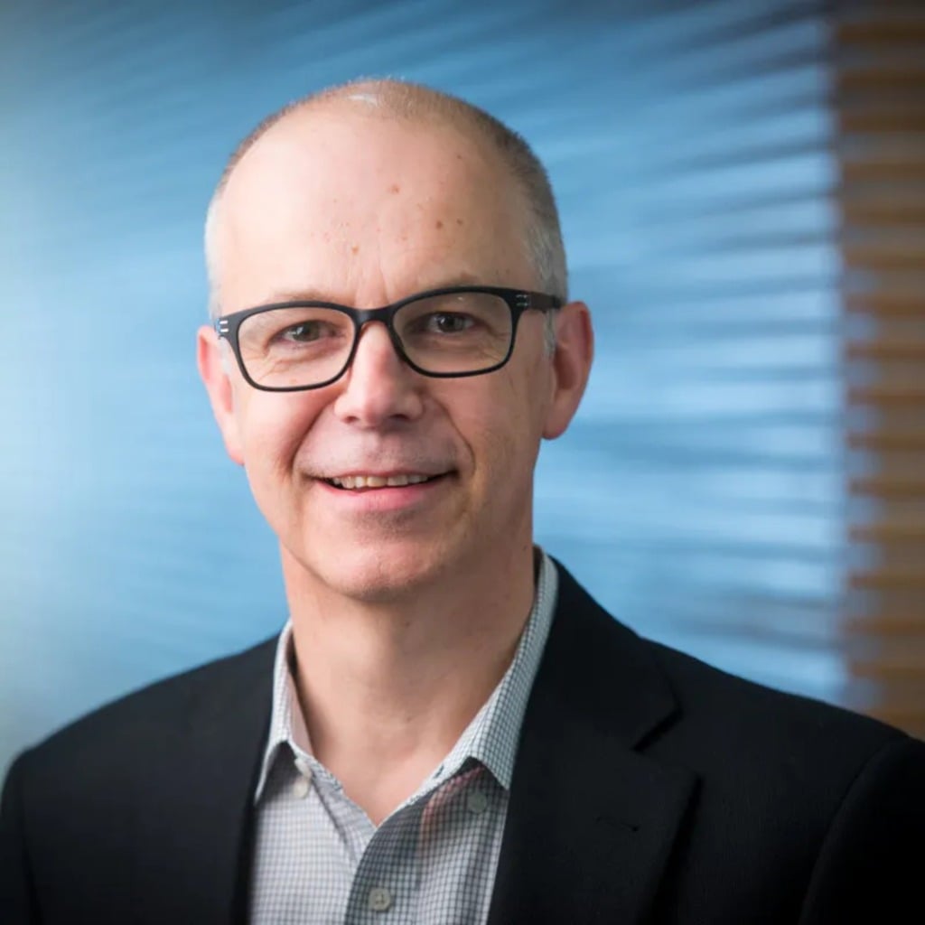 A smiling man with glasses in a black blazer, standing against a blue and wooden background, related to bowel cancer research.