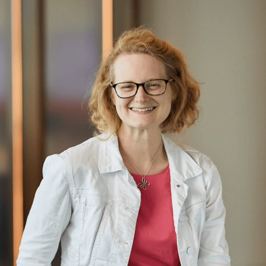 A smiling woman with curly hair, wearing glasses and a pink shirt, stands in a well-lit indoor setting.