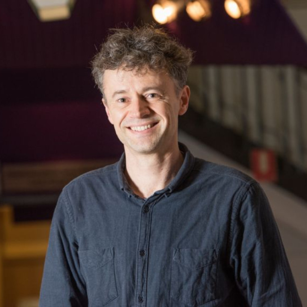 Smiling man with curly hair in a dark shirt, standing in a brightly lit space, related to early-onset bowel cancer centre of research excellence.