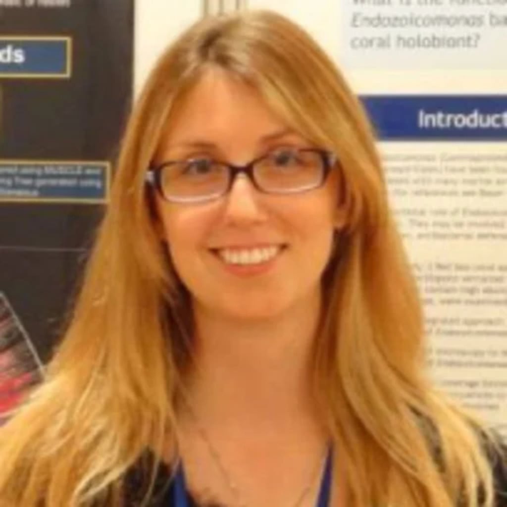A woman with long blonde hair and glasses smiles in front of a research poster related to early-onset bowel cancer.