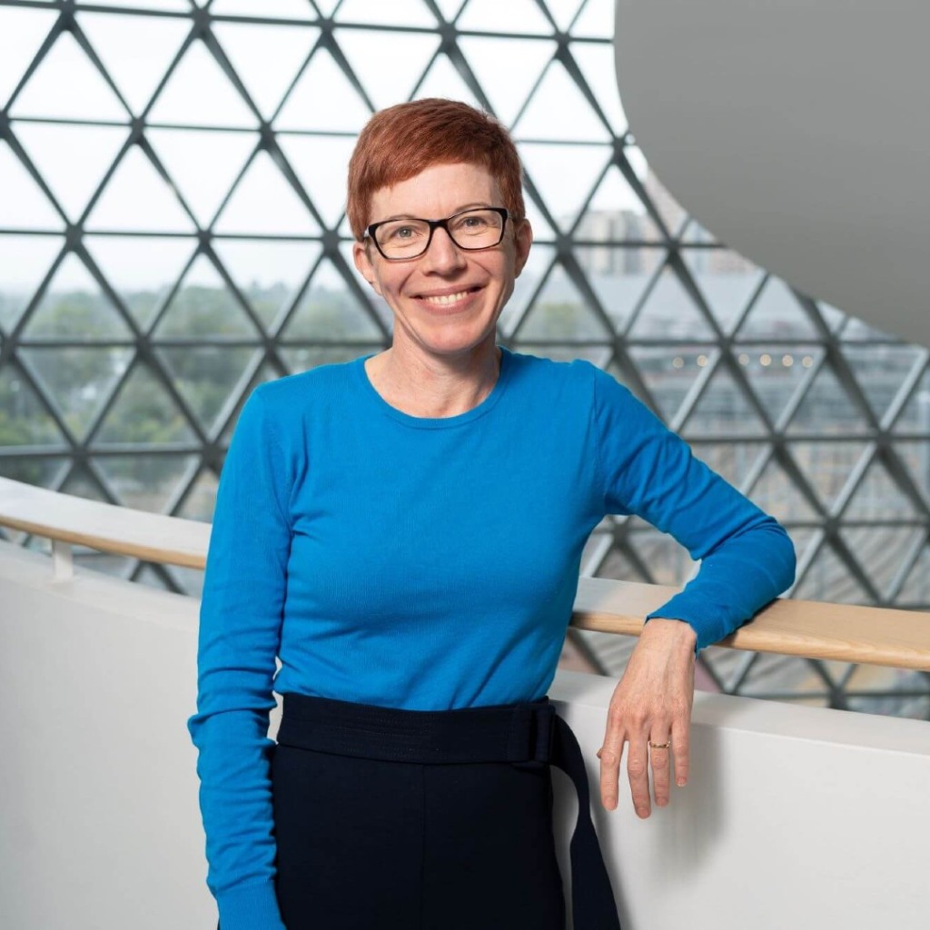 A woman in a blue long-sleeve shirt smiles while leaning on a railing, with a geometric glass backdrop.
