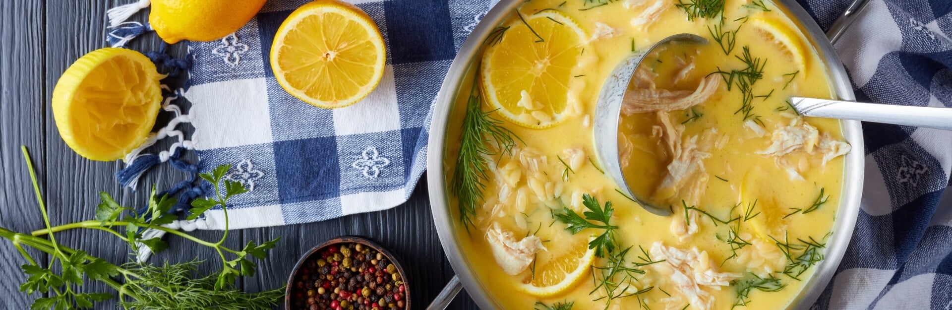 A pot of Avgolemono soup with lemon slices, shredded chicken, and fresh herbs on a checkered cloth and wooden surface.