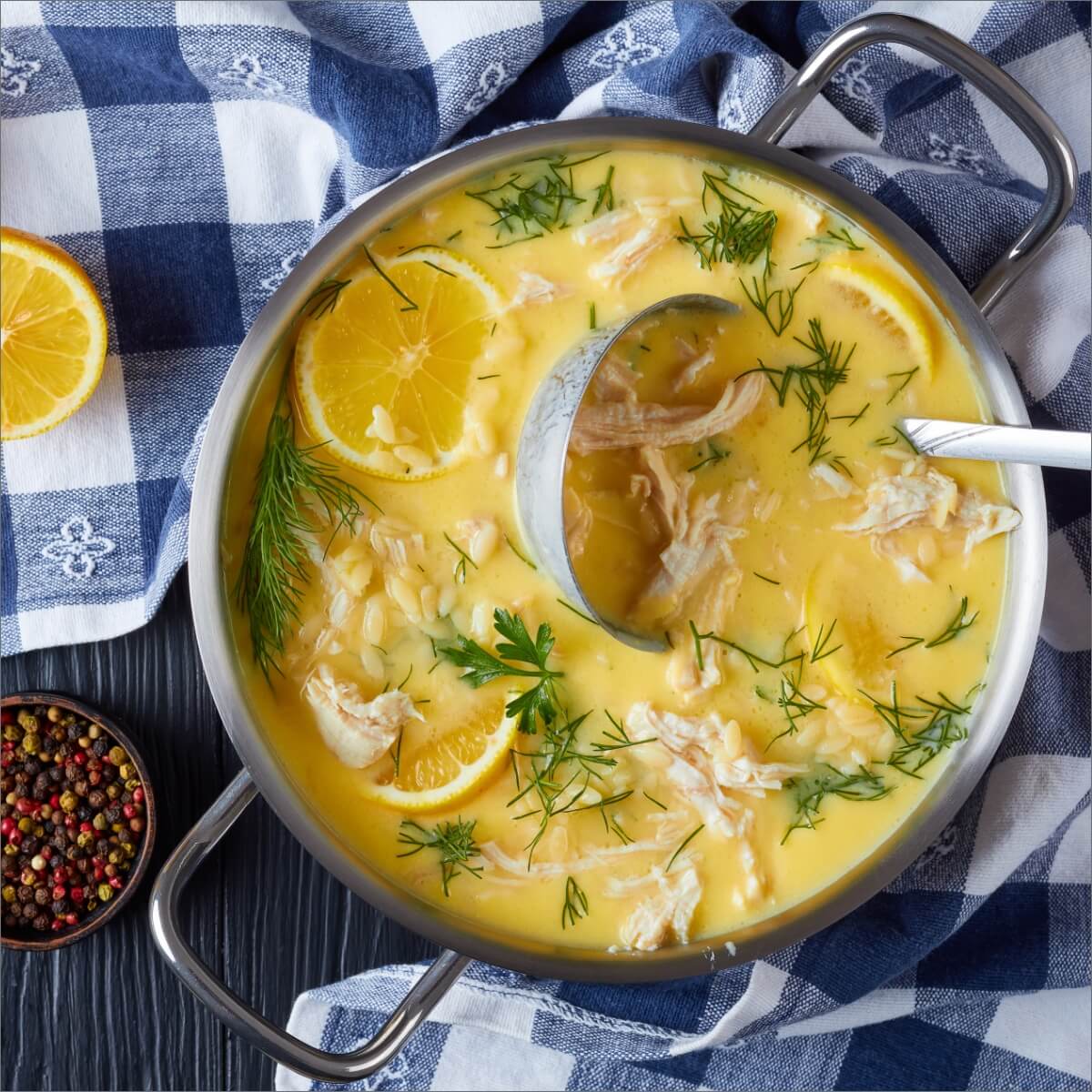 A pot of avgolemono soup with chicken, lemon slices, and herbs, next to a bowl of mixed peppercorns and a checkered cloth.