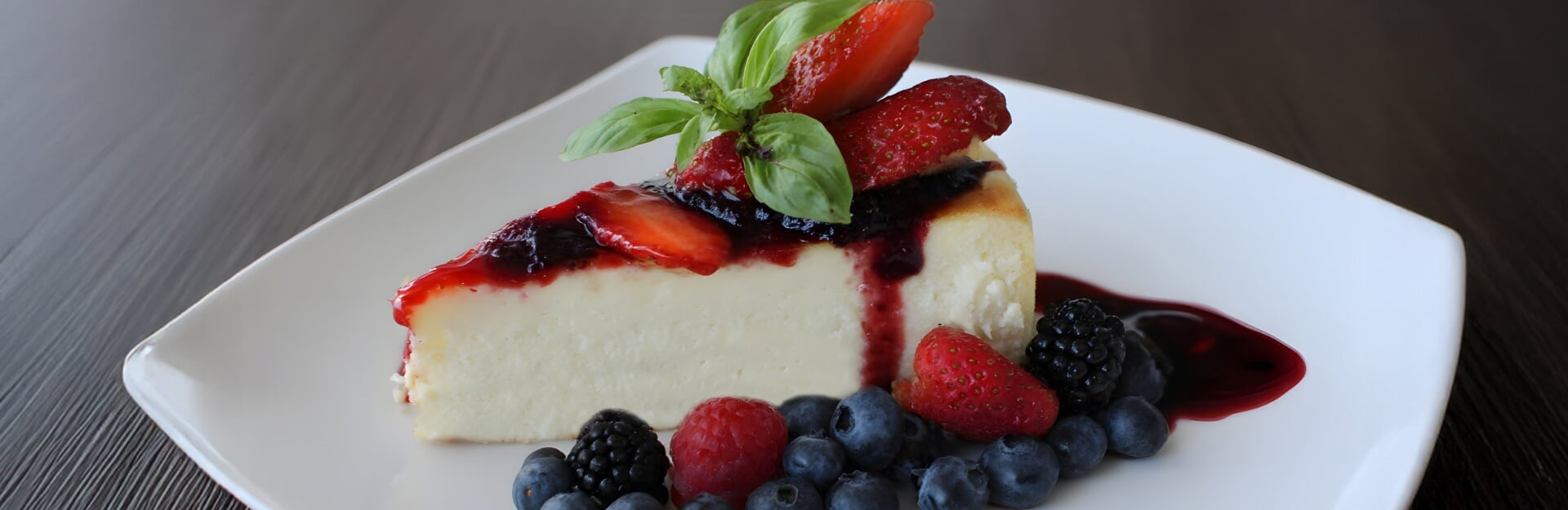 A slice of baked ricotta cake topped with strawberries, blueberries, and a berry sauce on a white plate.
