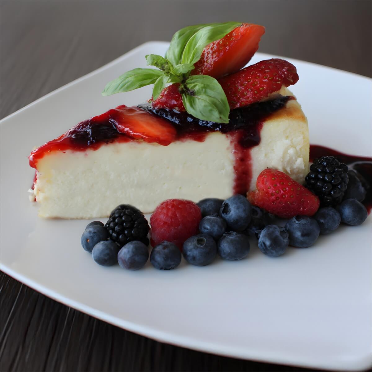 A slice of baked ricotta cake topped with strawberries, basil, and berry sauce, surrounded by fresh berries on a white plate.