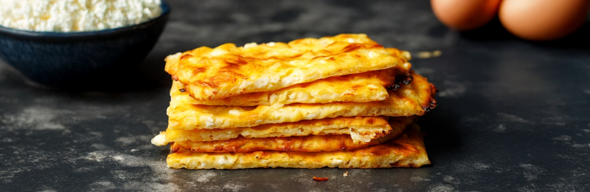 A stack of golden, crispy flatbreads beside a bowl of cottage cheese and eggs on a dark surface.