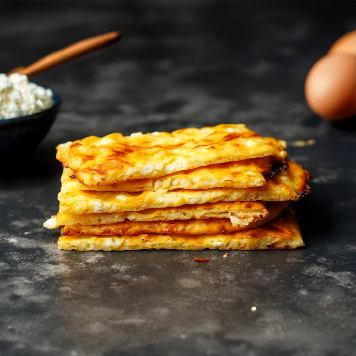 A stack of golden cottage cheese crackers on a dark surface, with a bowl of cottage cheese and an egg in the background.