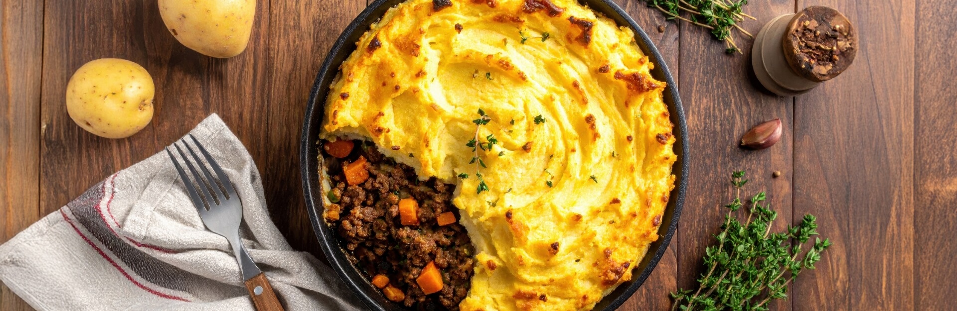 A shepherd's pie in a skillet, surrounded by potatoes, a fork, herbs, and a spice jar on a wooden surface.