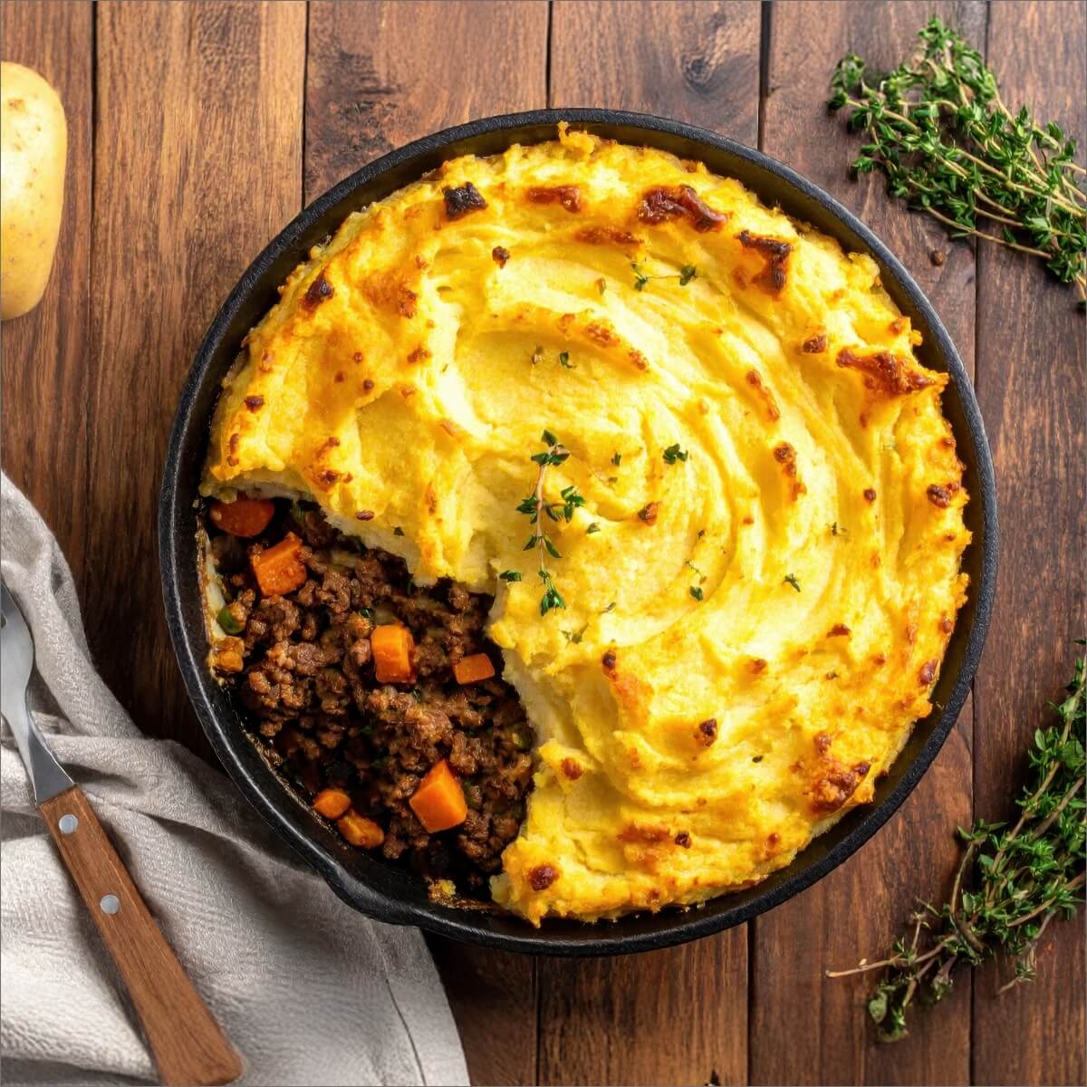 A cast iron dish of cottage pie with golden mashed potato topping and ground meat filling, garnished with thyme.