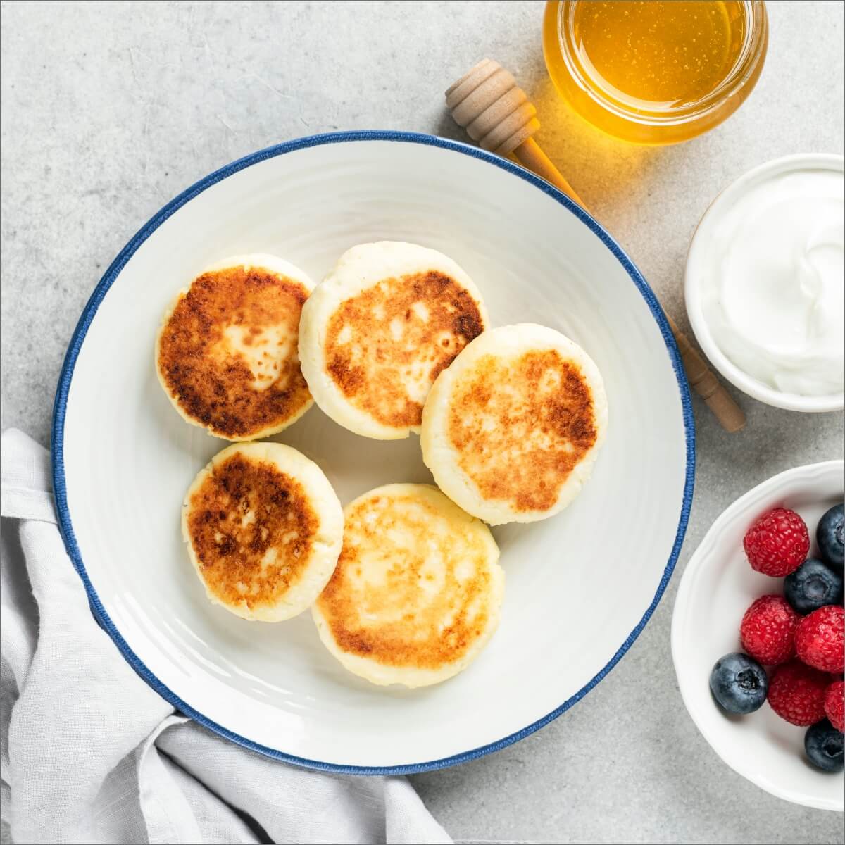 A bowl of mini banana pancake bites, honey, yogurt, and a dish of berries on a gray surface.
