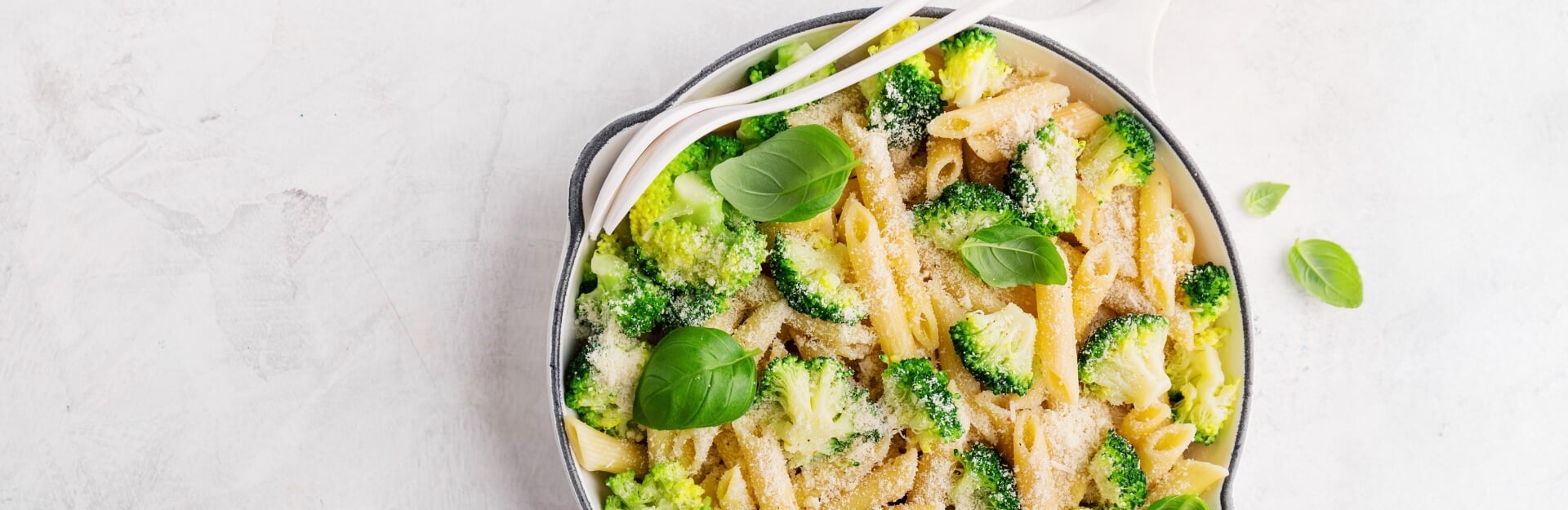 A bowl of pasta with broccoli, topped with grated cheese and fresh basil leaves, served with utensils.