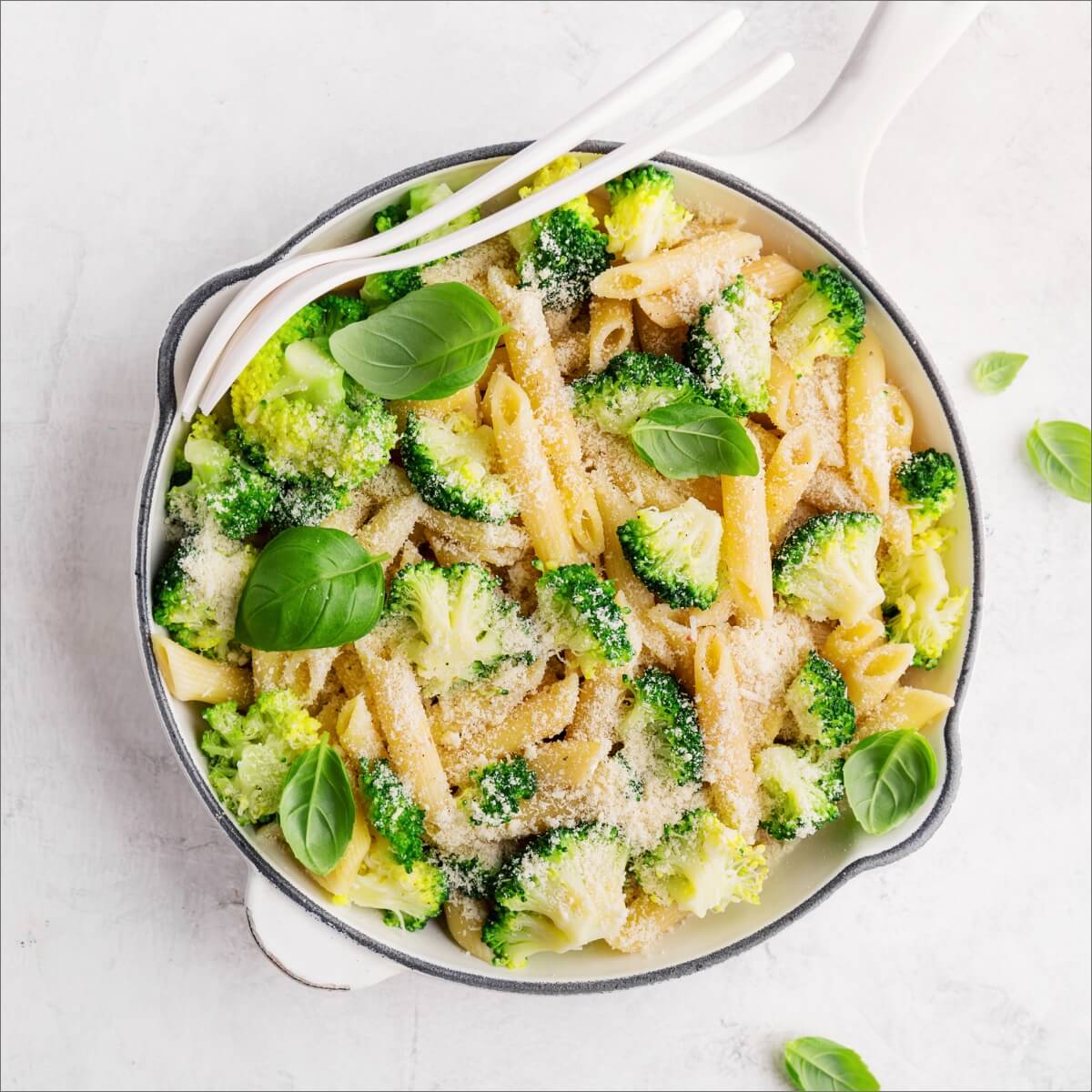 A bowl of pasta with broccoli, topped with grated cheese and fresh basil leaves, with two forks beside it.