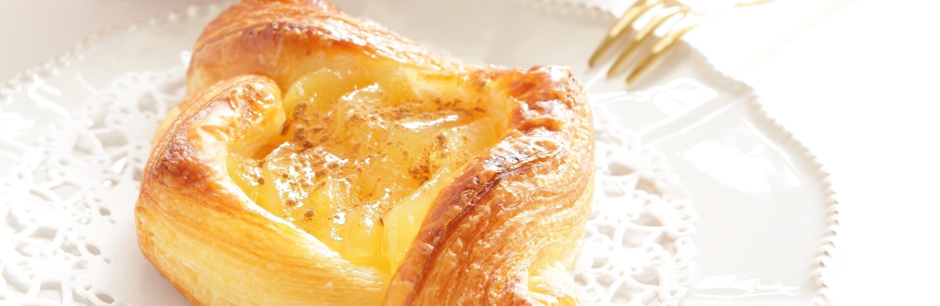 A golden-brown apple Danish pastry sits on a white plate with a lace doily.