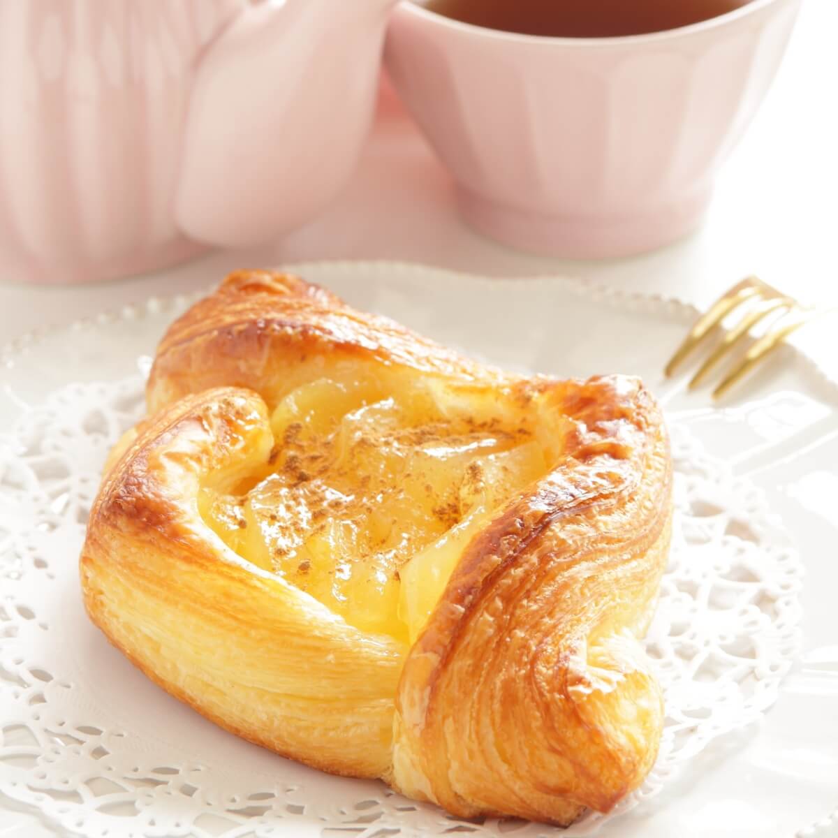 A golden-brown apple Danish pastry on a lace doily, with a pink teapot and cup in the background.