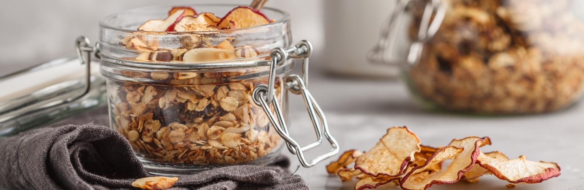 A glass jar filled with granola and dried fruit slices, next to a folded dark cloth and another jar in the background.
