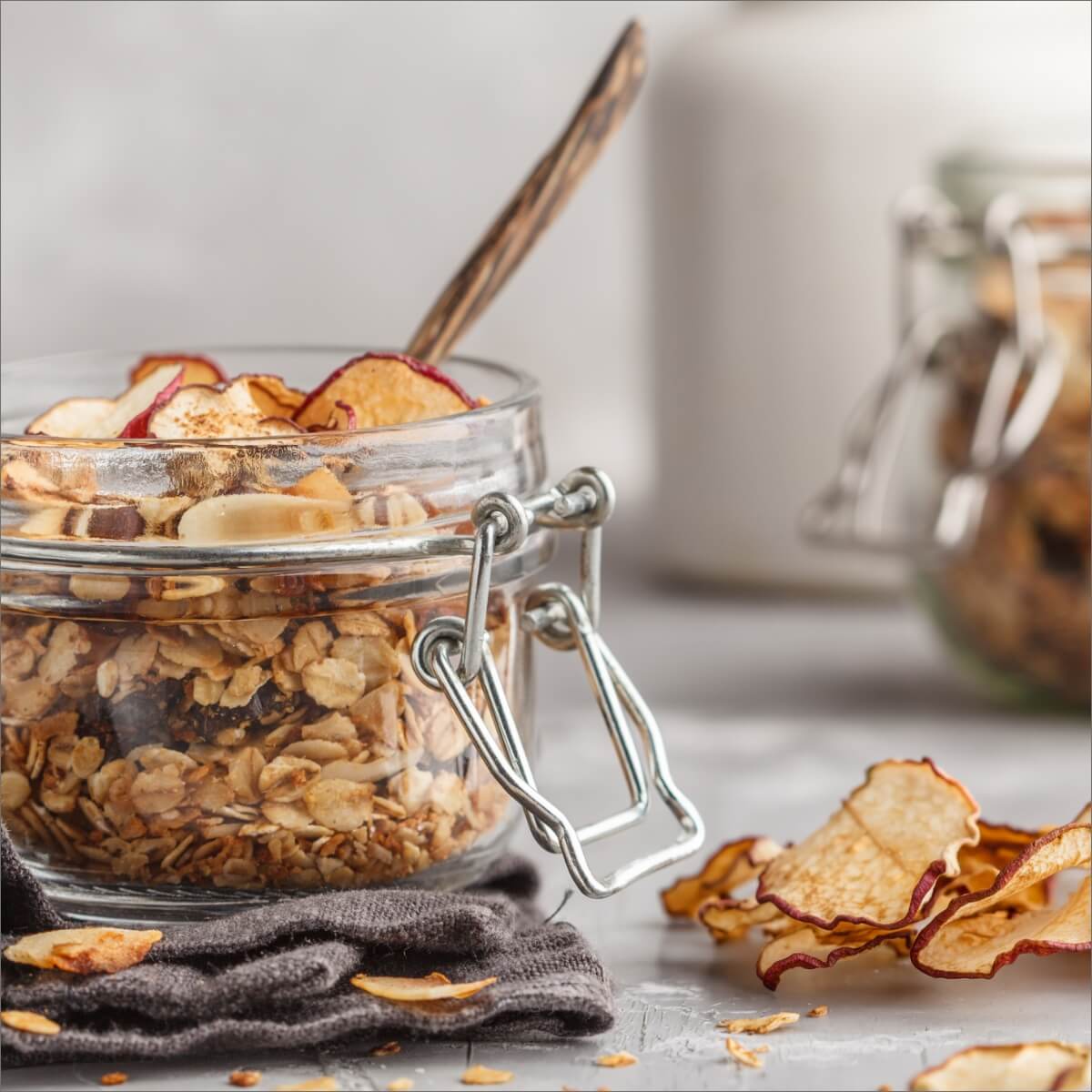 A glass jar of granola with apple slices, a wooden spoon, and scattered apple chips on a gray surface.