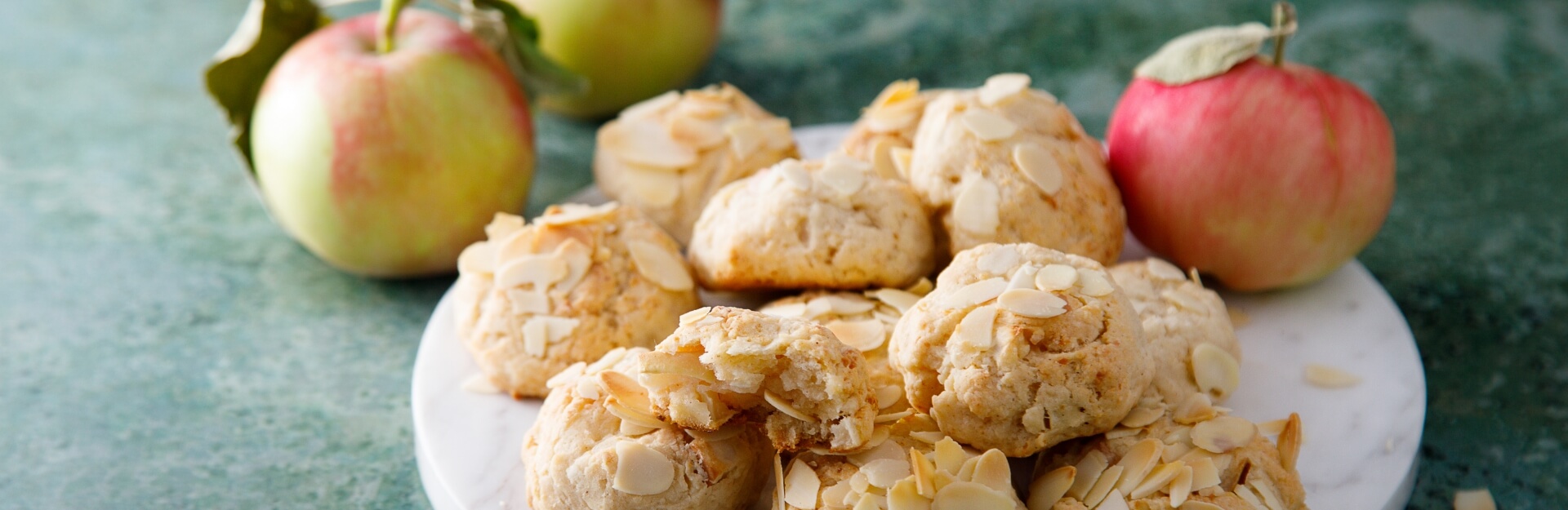 A plate of almond cookies with sliced almonds, surrounded by two whole apples on a green background.