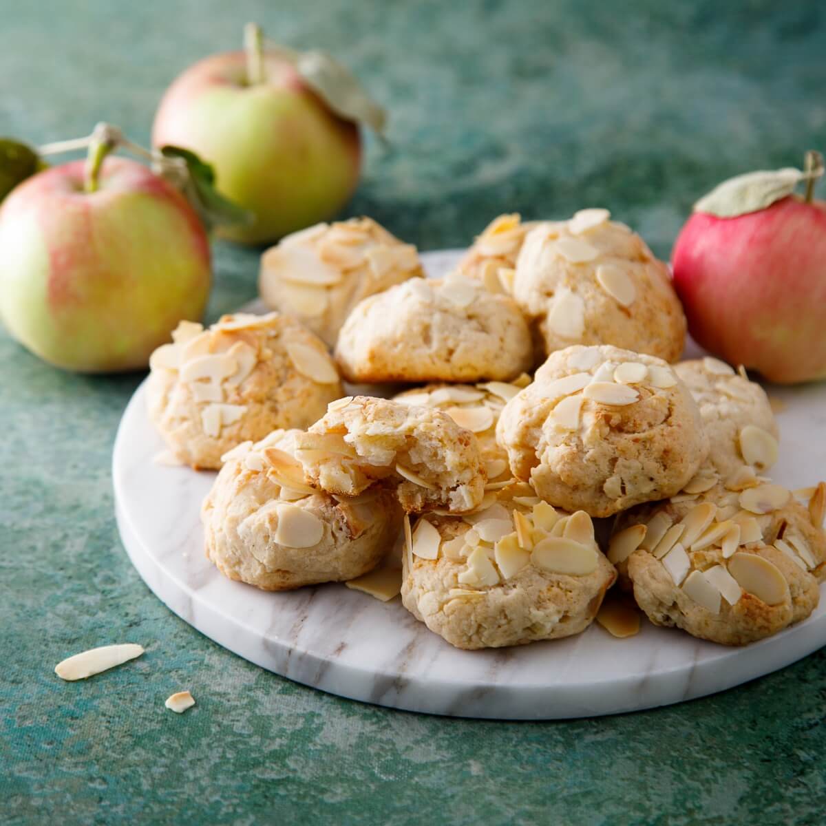 A plate of apple oat cookies topped with sliced almonds, surrounded by fresh apples on a green background.