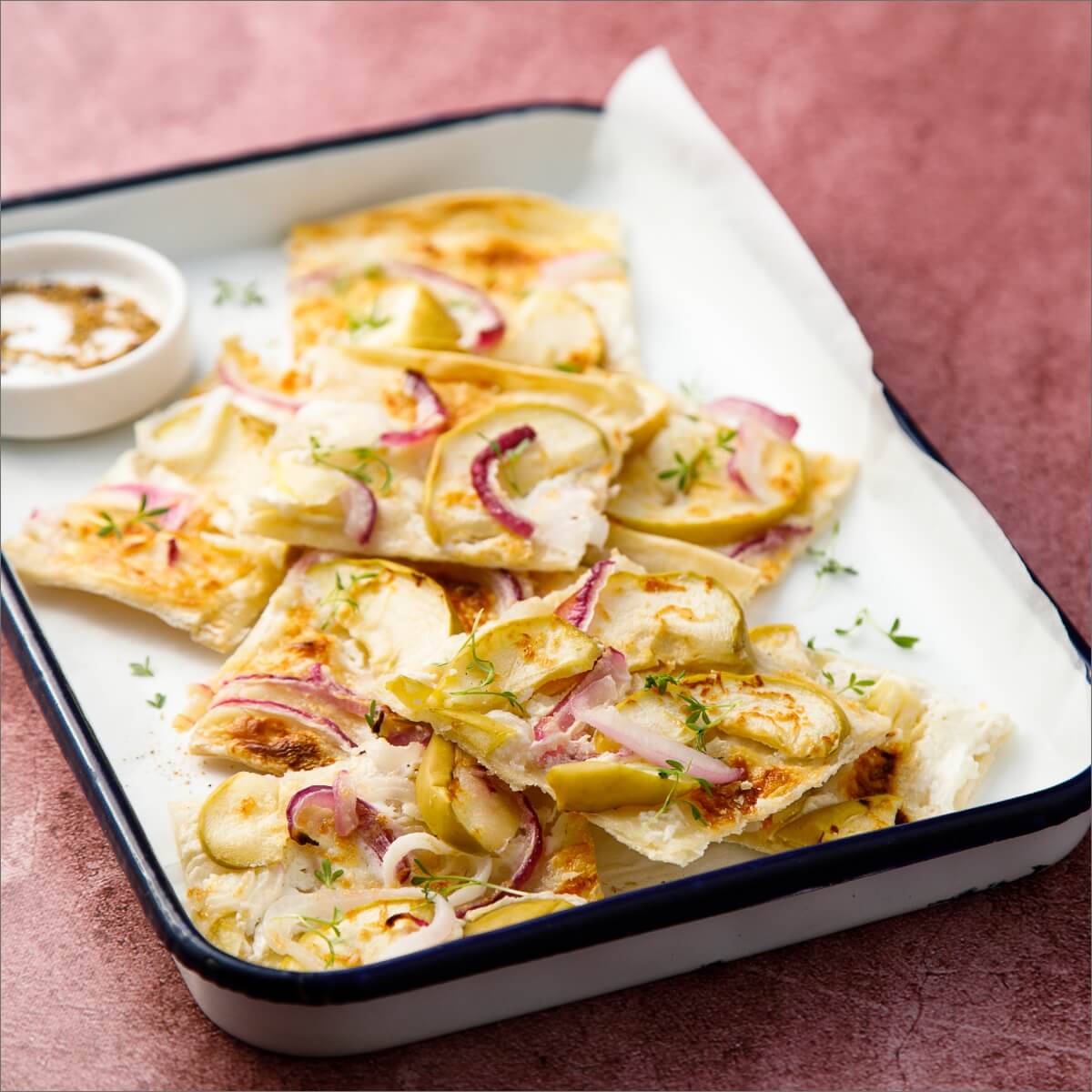A plate of apple, onion, and goat cheese tart slices garnished with herbs, accompanied by a small bowl of seasoning.