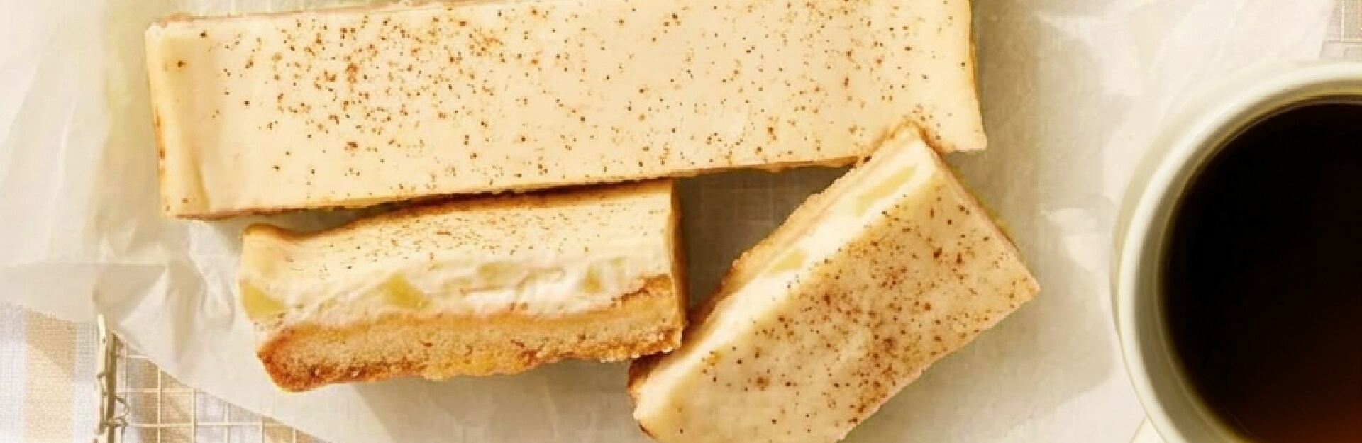 Three slices of apple and sour cream dessert on parchment paper, with a cup of dark coffee nearby.