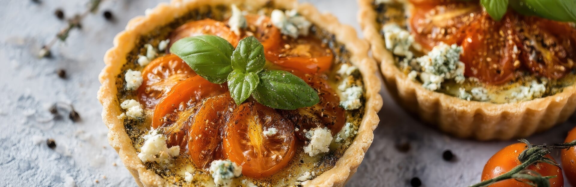 Two mini feta quiches topped with sliced tomatoes and basil, surrounded by fresh cherry tomatoes and herbs.