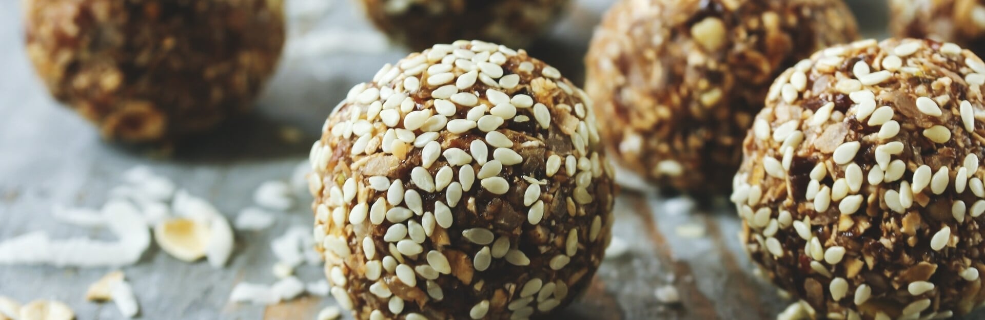 Close-up of protein balls coated in sesame seeds on a wooden surface, with a few scattered ingredients nearby.