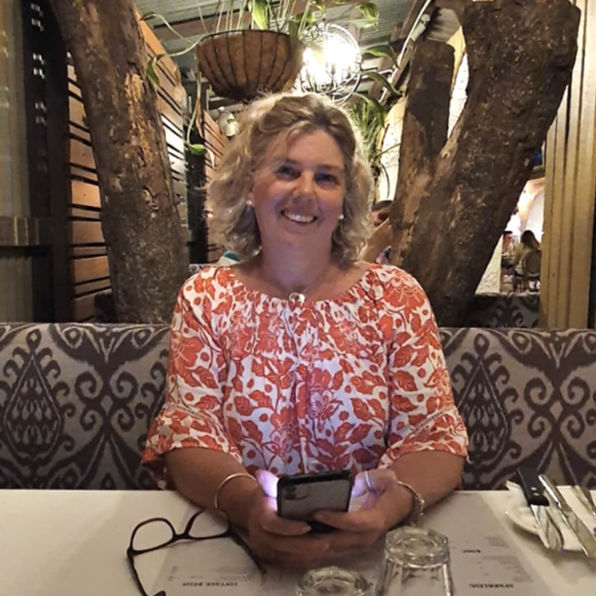 A smiling woman with curly hair sits at a table, holding a phone, in a cozy restaurant setting with wooden decor.