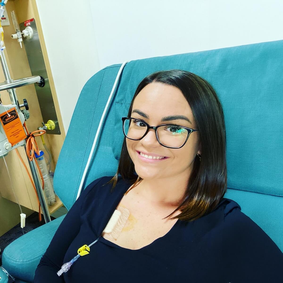 A woman with long hair and glasses smiles while sitting in a medical chair, with an IV line visible on her chest.