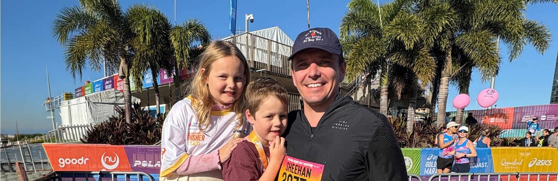 A man holds a boy while a girl stands beside them, smiling at a marathon event with palm trees in the background.