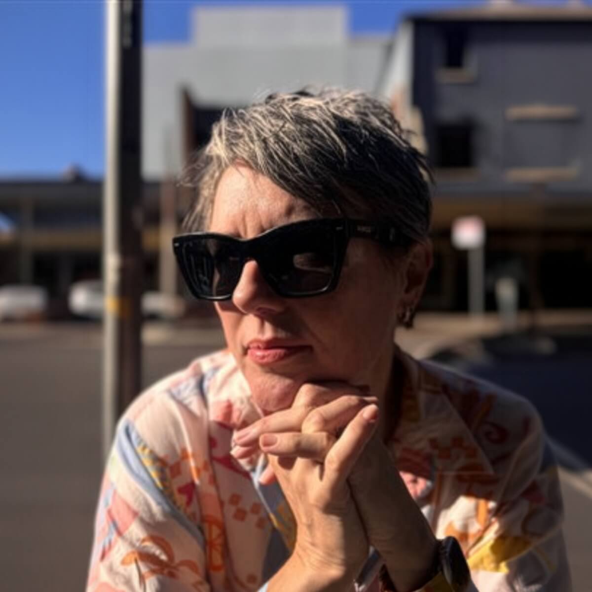 A woman with short gray hair wears sunglasses and a colorful shirt, resting her chin on clasped hands outdoors.